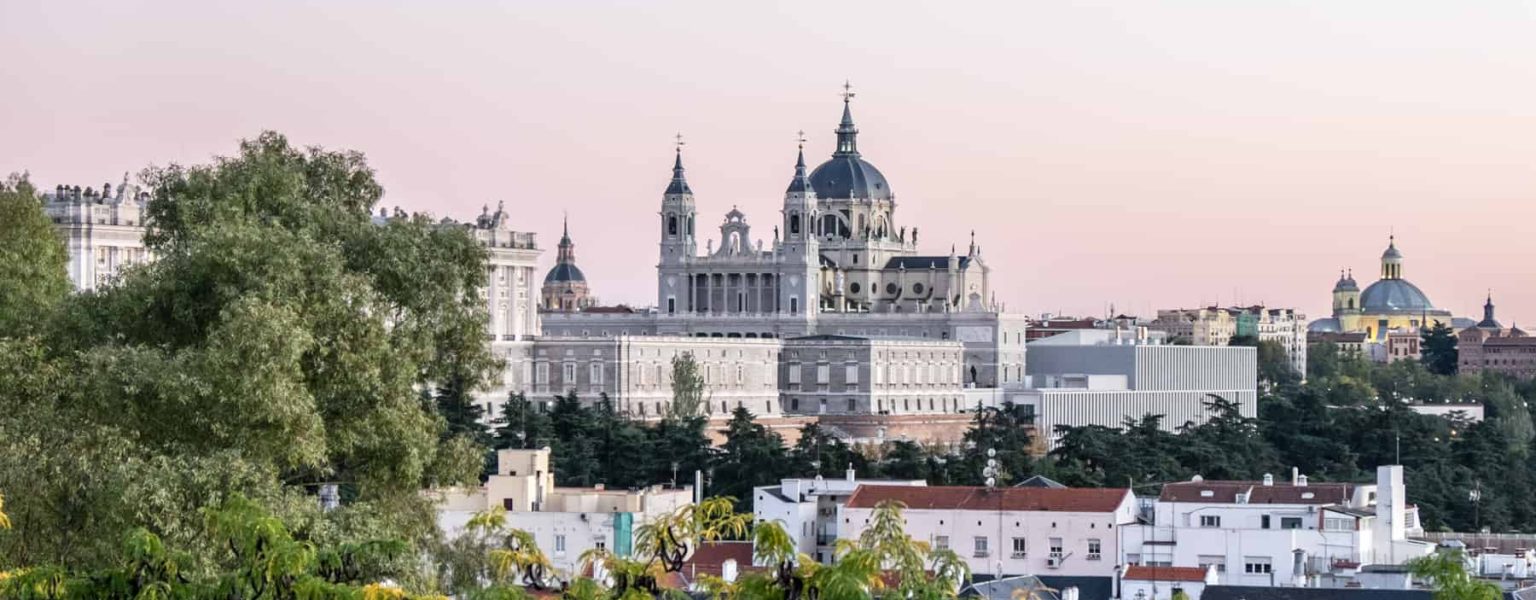 A silvery white palace complex with dark grey dome roof and spires is elevated about square city buildings in Madrid. The view is from within the foliage of parkland, and at a distance.