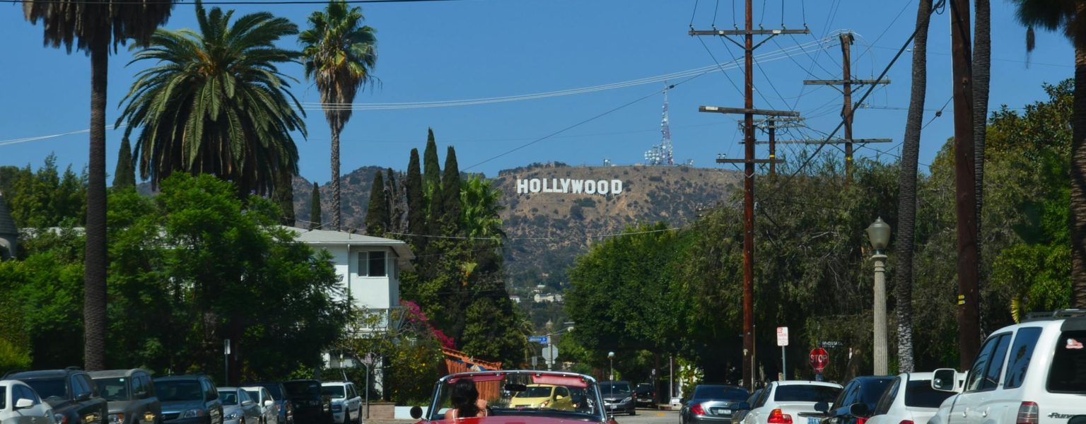 Red convertible driving down a sunny street towards the iconic Hollywood sign in Los Angeles.